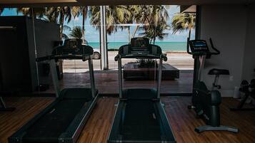 Un gimnasio con cintas de correr y una máquina elíptica, con vistas a una playa con palmeras.