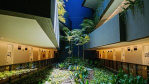 Un pasillo de hotel con puertas numeradas, un jardín central con plantas y un techo azul.