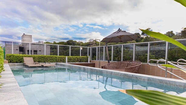 A swimming pool with lounge chairs and an umbrella, surrounded by a glass fence and greenery. - Vivendas #404 - Apto em Cabo Branco by Carpediem (João Pessoa)