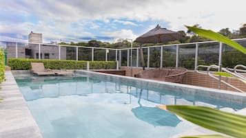Piscina con sillas de descanso y una sombrilla, rodeada por una valla de vidrio y follaje.