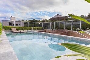 A swimming pool with lounge chairs and an umbrella, surrounded by a glass fence and greenery. - Vivendas #404 - Apto em Cabo Branco by Carpediem (João Pessoa)