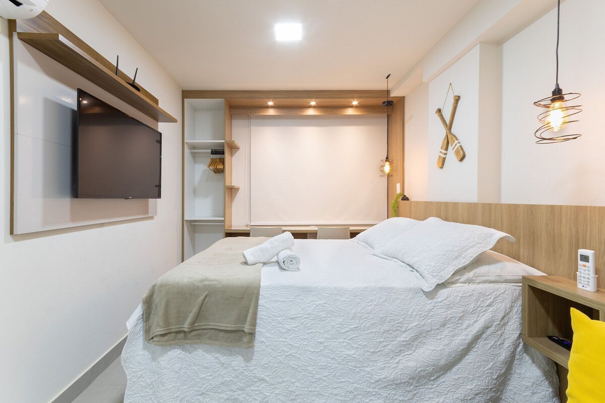 A neatly made bed with a white comforter and a beige throw blanket, a bedside table with a lamp, and a mounted TV on the wall.