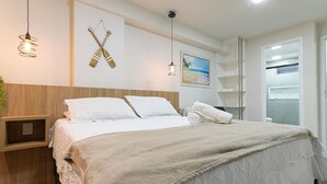 A neatly made bed with a fluffy blanket, a wooden headboard, and a wall-mounted shelf with a small plant. - Vivendas #404 - Apto em Cabo Branco by Carpediem (João Pessoa)