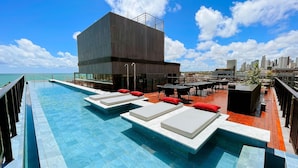 A rooftop pool with lounge chairs and a bar area, overlooking a cityscape. - Acqua Tambaú Prime - By Almare (João Pessoa)
