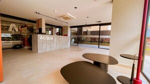 A reception area with a marble counter labeled 'Acqua,' a glass door leading outside, and a seating area with a table and chairs. - Acqua Tambaú Prime - By Almare (João Pessoa)