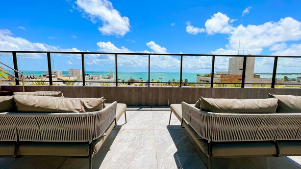 A terrace with wicker furniture, a view of the ocean, and buildings in the background. - Acqua Tambaú Prime - By Almare (João Pessoa)