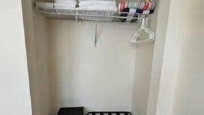 A small, narrow closet with a shelf holding folded towels and a hanging rack with clothes. There is a small storage unit with drawers and a black and white striped chair. - Cozy Studio – Free Bikes – 1 Mi to Beach (Bonita Springs)