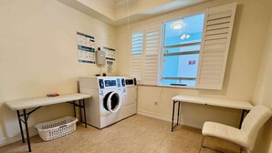 A laundry room with a washing machine, two tables, a basket, and a window with blinds. - Cozy Studio – Free Bikes – 1 Mi to Beach (Bonita Springs)
