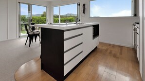 Et moderne køkken med en stor ø, et spiseområde med stole, og store vinduer med udsigt til grønt og bakker.