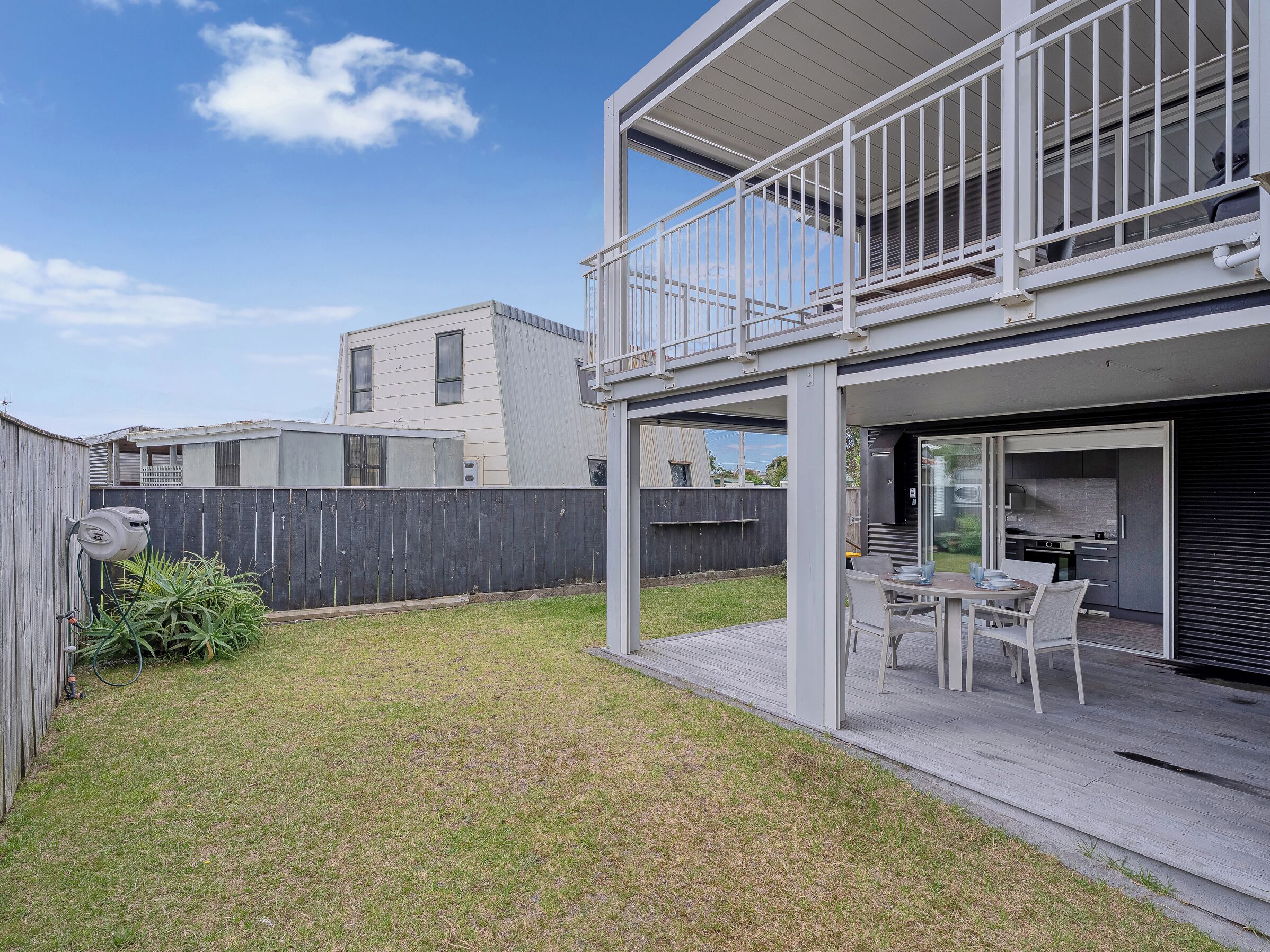 A modern two-story house with a balcony, a patio with outdoor furniture, and a fenced backyard.