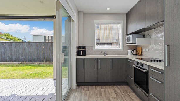 A modern kitchen with grey cabinets, a built-in microwave, and a window with blinds. - Soleil Studio - Whangamata Self-contained Unit (Whangamata)