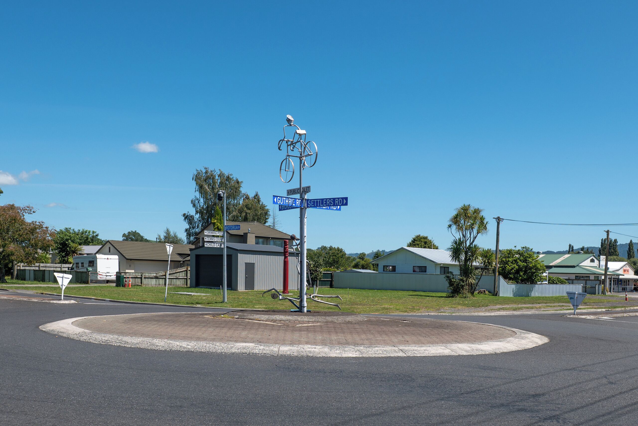 A roundabout with a bicycle sculpture at the center, surrounded by residential buildings and a clear blue sky.