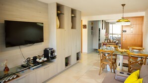 A modern living room with a flat-screen TV, a coffee machine, and a dining area with a glass table and wooden chairs. - Paraiso do Atlantico #102d - Style by Carpediem (Cabedelo)