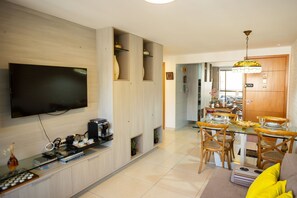 Ein modernes Wohnzimmer mit Flachbildfernseher, einer Kaffeemaschine und einem Essbereich mit Glastisch und Holzstühlen.