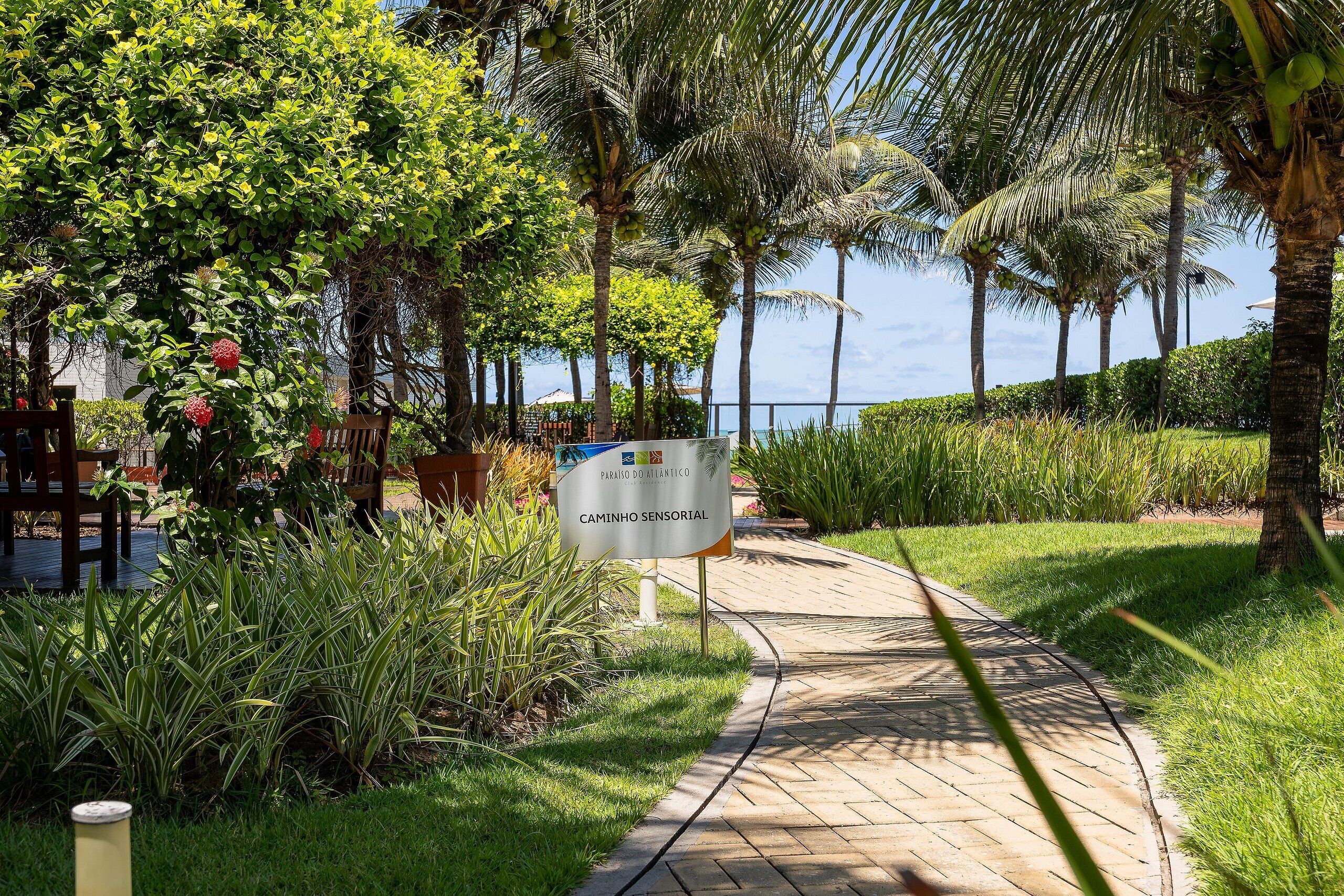 A paved pathway with a sign reading "CAMINHO SENSORIAL" surrounded by lush greenery and palm trees.