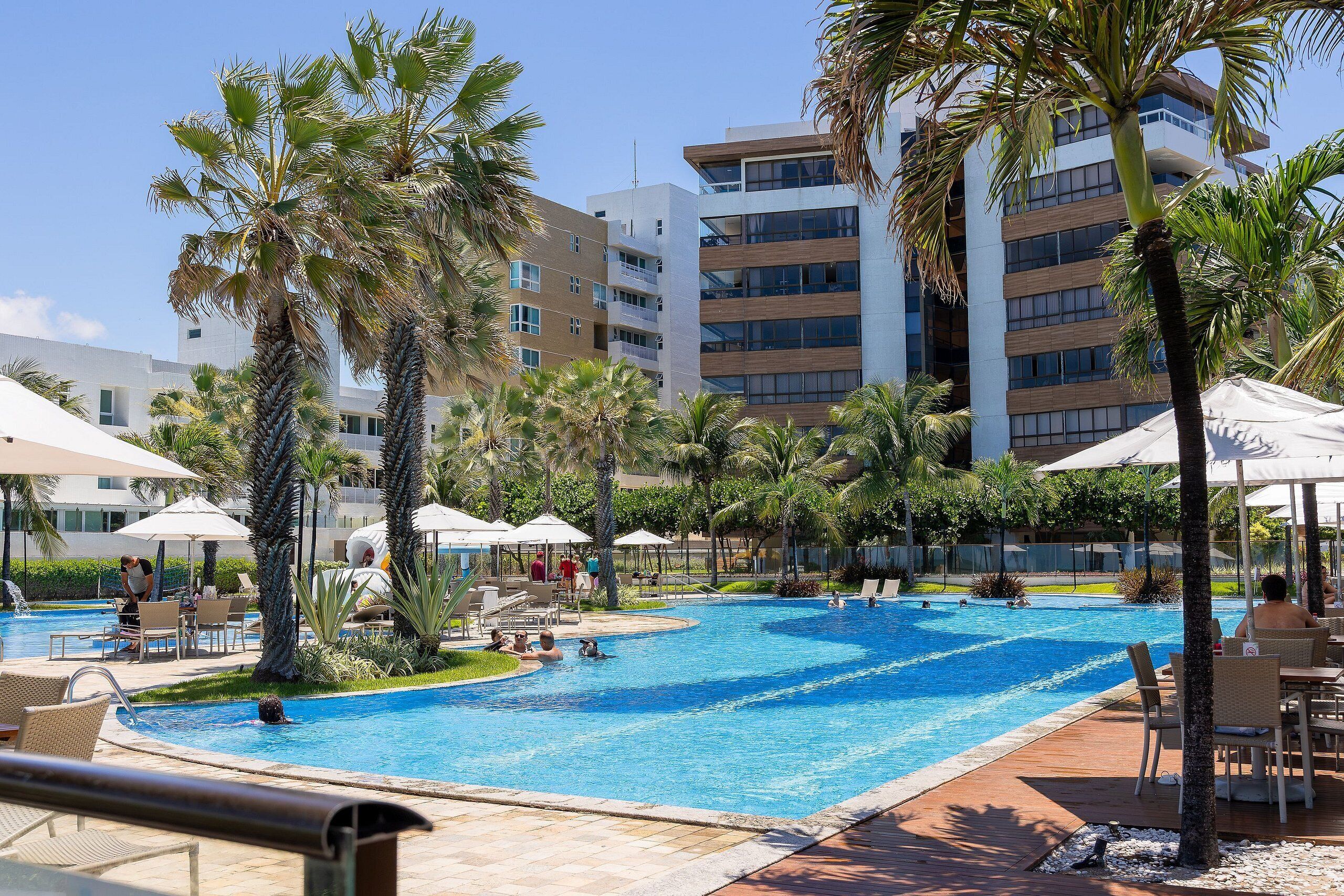 A swimming pool with lounge chairs and umbrellas, surrounded by palm trees and modern buildings.