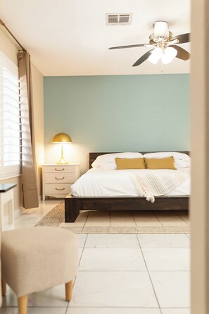 Une chambre à coucher comprenant un lit, une table de chevet, une lampe, un ventilateur de plafond et une chaise.