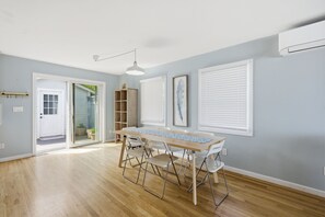 Une pièce avec un plancher en bois, une table à manger avec des chaises, une porte blanche et une fenêtre avec des stores.