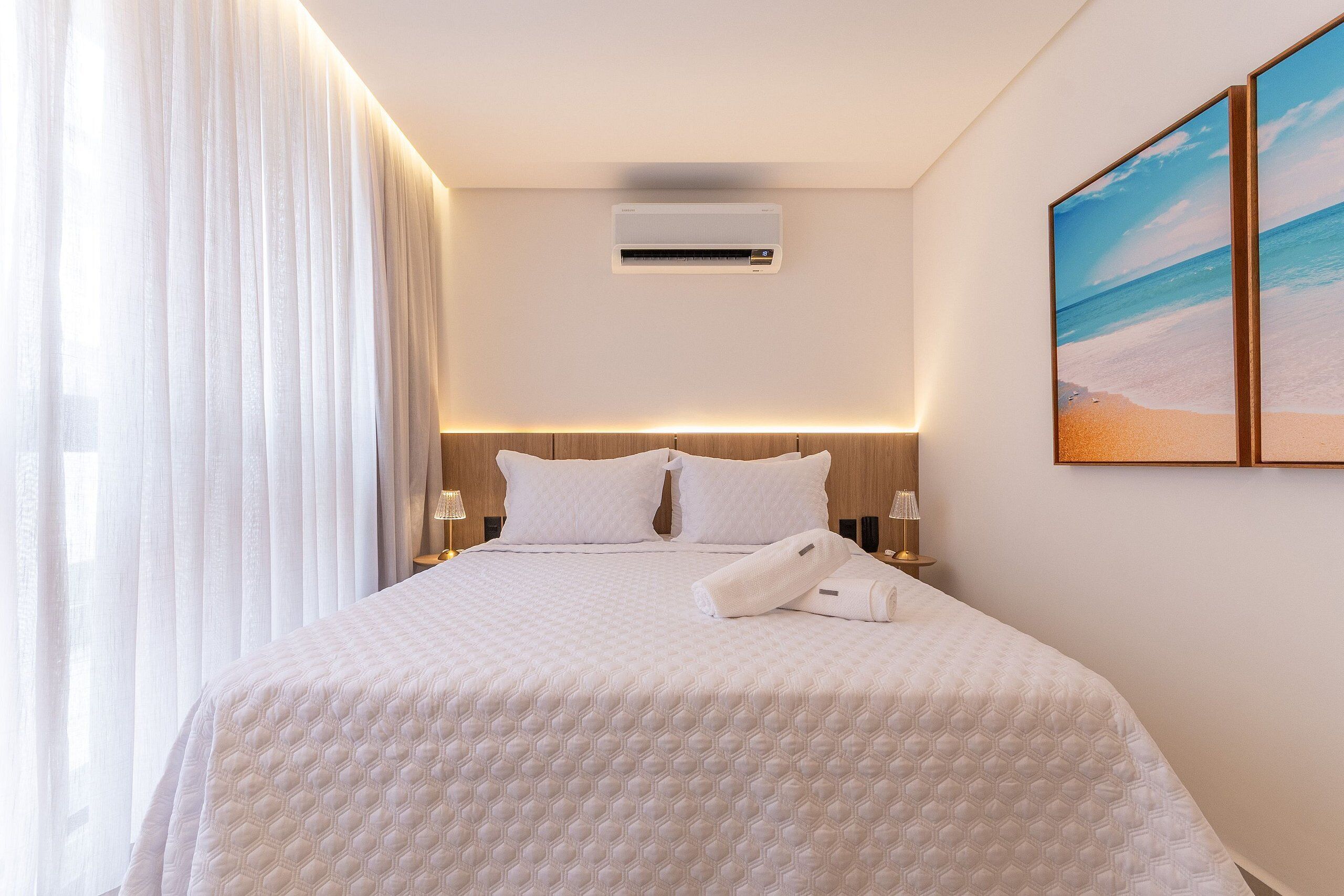 A hotel room with a large bed, white bedding, an air conditioning unit, a nightstand with a lamp, and a beach-themed painting on the wall.