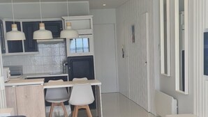 Un saló modern amb un sofà, una zona de menjador amb taula i cadires, una cuina amb armaris i un taulell, i un passadís amb mirall.