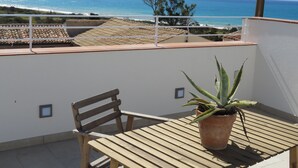 Een balkon met een houten tafel en een bloempot, met uitzicht op de kust onder een heldere lucht.