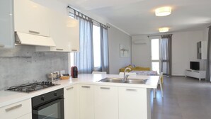Een moderne keuken met witte kasten, een gasfornuis en een inbouwoven. Er is een eethoek met een gele tafel en stoelen, en een televisie op een standaard.