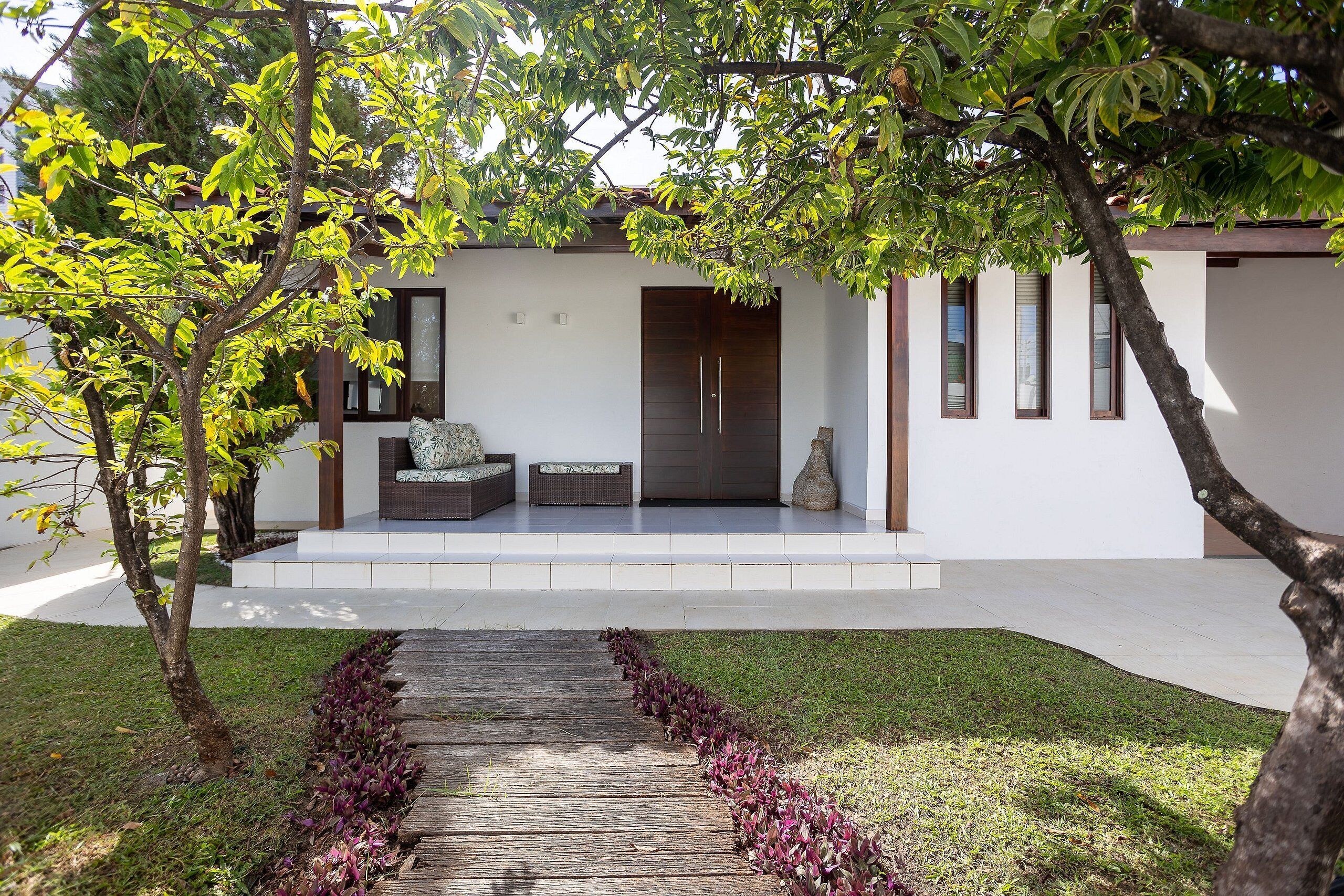 A modern house with a wooden deck leading to a front door, surrounded by greenery and a well-maintained lawn.
