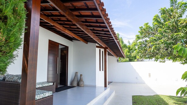 Ein überdachter Außenbereich mit einer Holnpergola, einem Korbsessel mit Kissen und zwei Steinvasen.