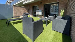 Un patio al aire libre con mobiliario de mimbre, una pared de ladrillo y una puerta corrediza de cristal que da a un espacio interior.