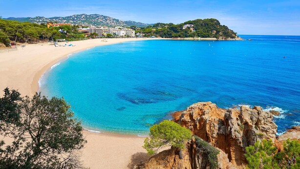 Una playa costera con aguas azules cristalinas, acantilados rocosos y un pueblo al fondo.