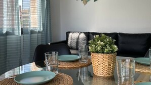 Un coin repas avec une table en verre, des sets de table en fibres tressées et des assiettes de couleur turquoise. On y trouve également un panier tressé contenant des plantes et une œuvre d’art murale au-dessus de la table.