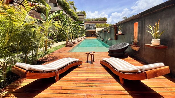 A wooden deck with two lounge chairs and a small table, leading to a swimming pool surrounded by lush greenery and a modern building. - Private and Peaceful Apartment 1BD 1BR (Tulum)