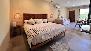 A spacious bedroom with a large bed, a wooden headboard, and two bedside tables. There is a sofa and a dining table in the adjacent living area. - Private and Peaceful Apartment 1BD 1BR (Tulum)