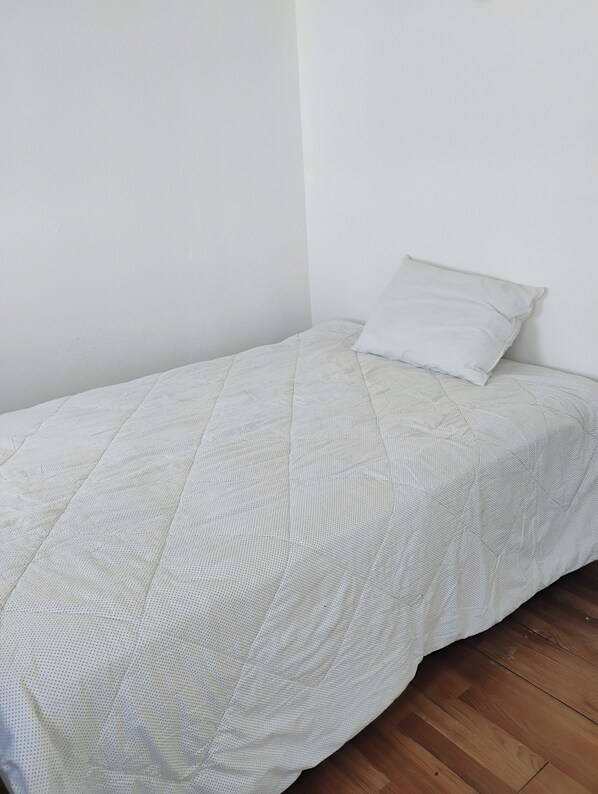 A small, neatly made bed with a quilted white bedspread and a single white pillow against a plain white wall.