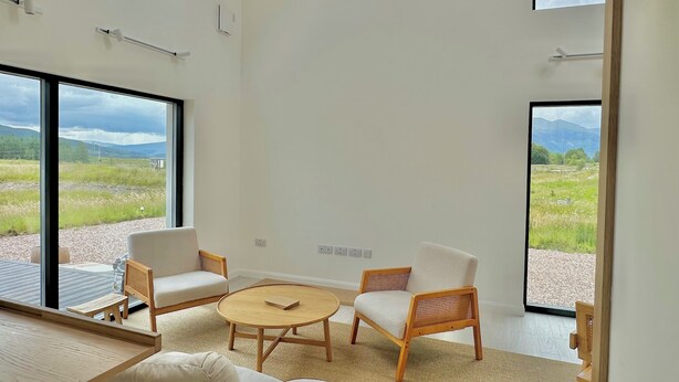 Una habitación amplia con ventanas grandes, una mesa redonda de madera, dos sillas de mimbre y un sofá.