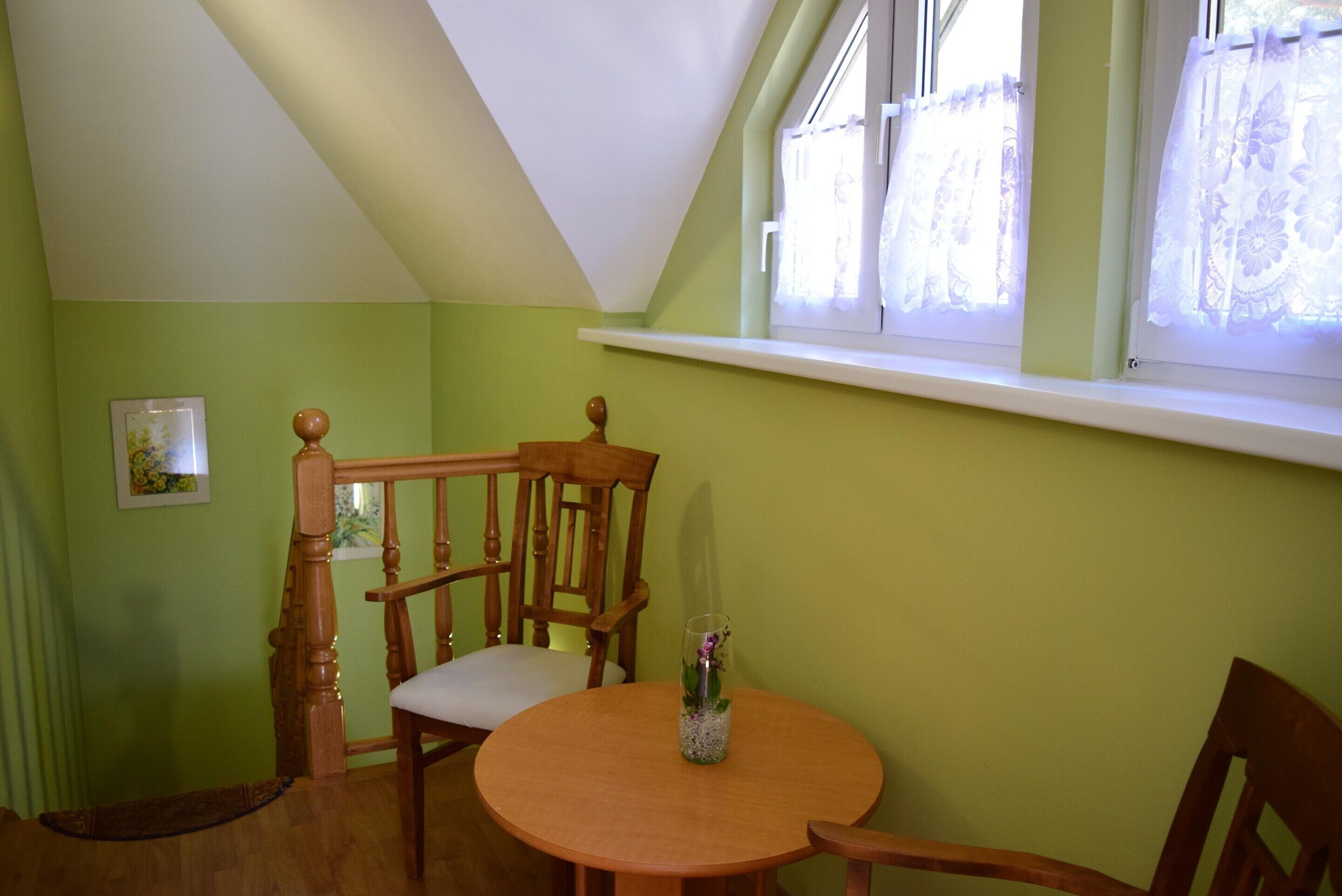 A room with a wooden dining table, chairs, a wooden chair with a cushioned seat, a small vase with flowers, and a window with lace curtains.