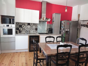 A modern kitchen with white cabinets, a red accent wall, a dining table with chairs, and a built-in oven. - CHAMBRECOSY town house between train station and historic center (Quimper)