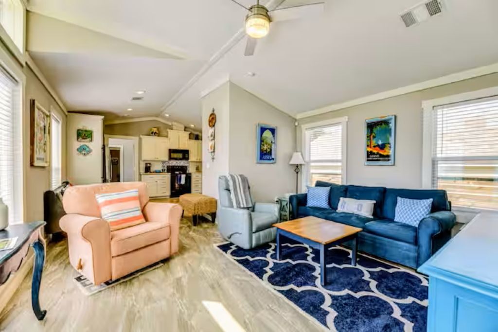 A living room with a sofa, armchair, and coffee table, a kitchen in the background, and a ceiling fan.
