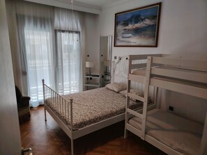 Ein Schlafzimmer mit Etagenbett, Nachttisch mit Lampe, Fenster mit transparenten Vorhängen und einem Bild an der Wand.