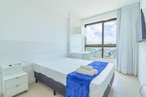 A modern hotel room with a large bed, a bedside table, a small desk, and a window offering a city view. - Parque Shopping #706 - Apt in Pina by Carpediem (Recife)