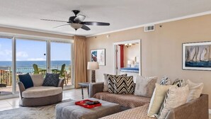 A living room with a sofa, ottoman, and a chair, a ceiling fan, and a large window with a view of the ocean.
