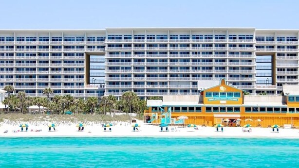 A large hotel with multiple floors and balconies, situated by a beach with clear turquoise water.