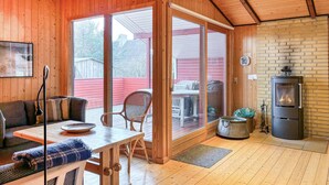 Ein gemütliches Blockhaus-Interieur mit Holzboden, einem Esstisch mit Stühlen, einer Couch, einem Kamin und einer großen Glasschiebetür, die auf eine Terrasse im Freien führt.