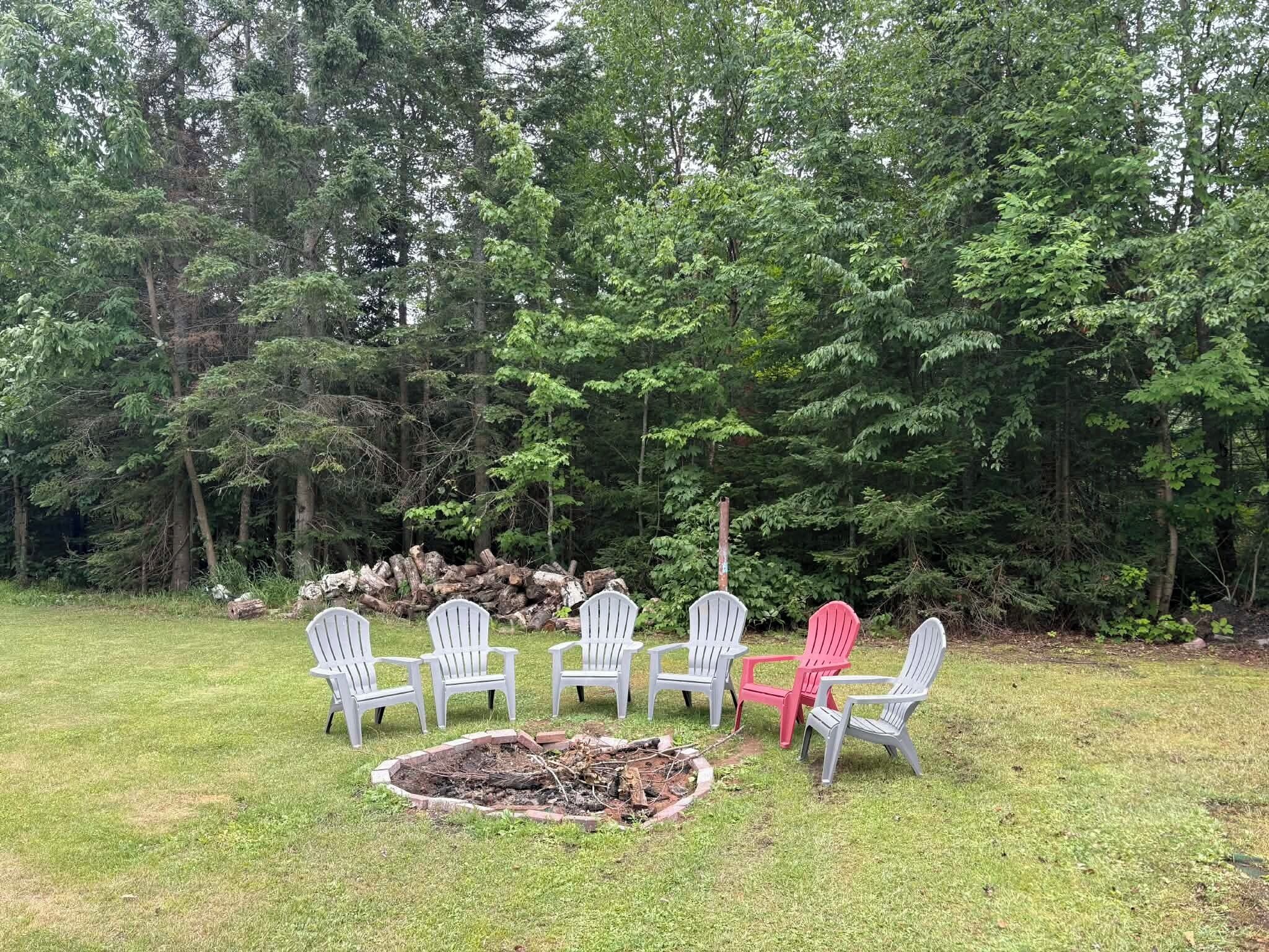 A grassy outdoor area with a fire pit and six Adirondack chairs, surrounded by dense trees.