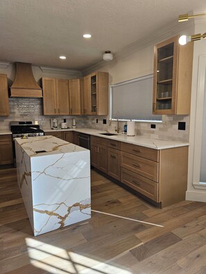 A modern kitchen with wooden cabinets, a marble island, and a built-in oven. - City Retreat (Sugar Land)