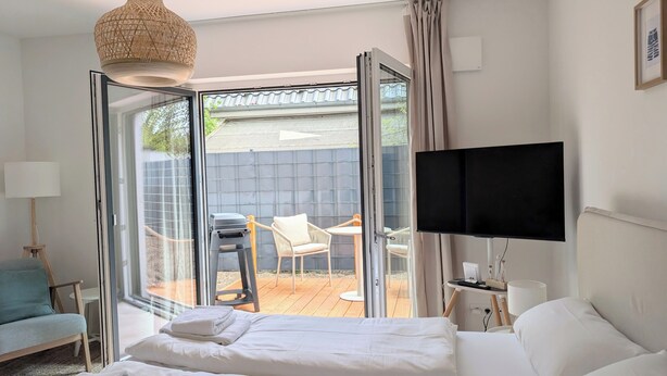 Ein Hotelzimmer mit Bett, Fernseher, Schreibtisch mit Computer, Stuhl und Blick ins Freie.