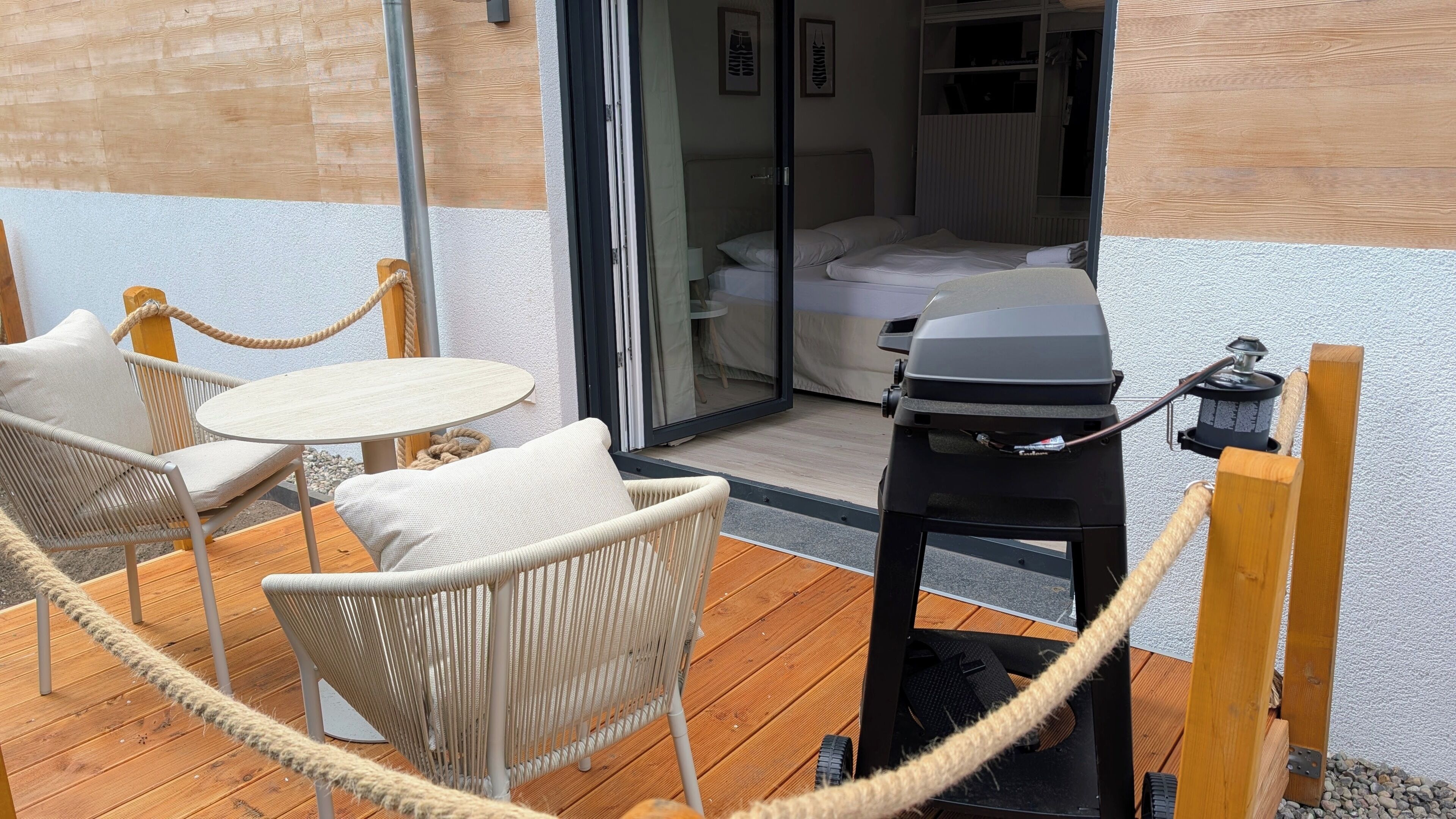 A patio with wicker furniture, a grill, and a view into a bedroom with a bed and a wardrobe.