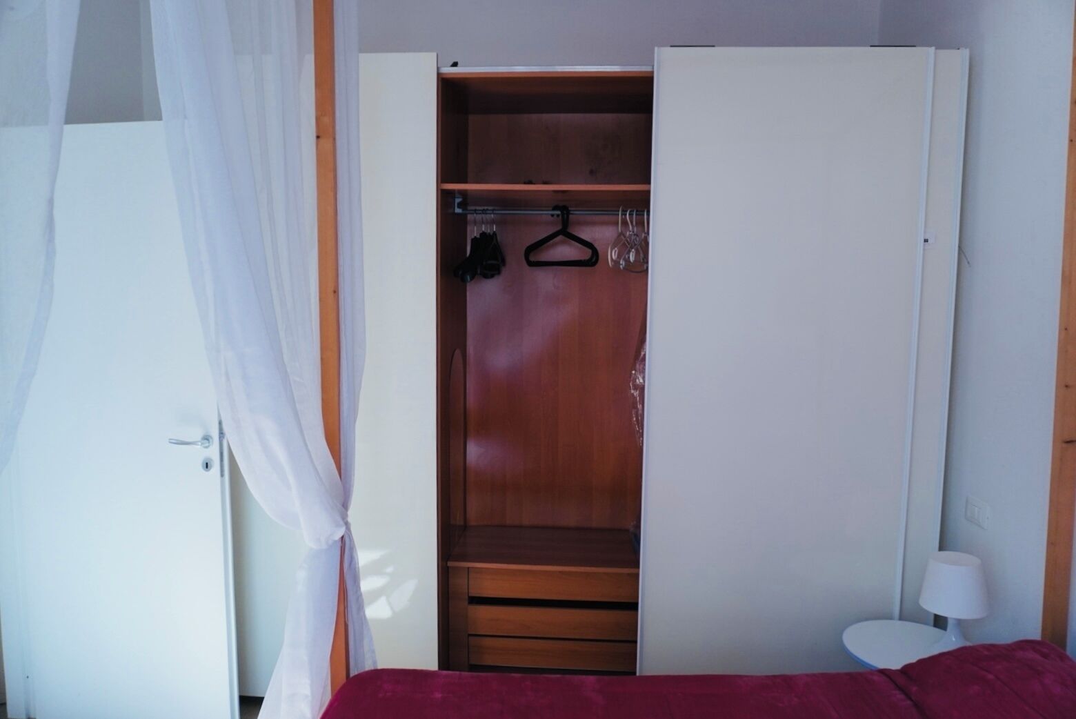 A bedroom with a wooden wardrobe, a bed with a red cover, a bedside table with a lamp, and a hanging coat rack.