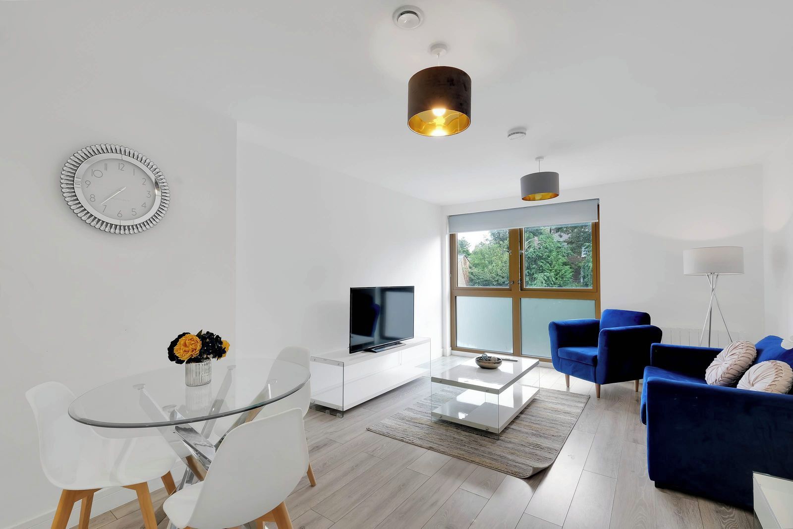 Ein modernes Wohnzimmer mit einem Glastisch, einem Flachbildfernseher, einem blauen Sofa und einem großen Fenster mit Blick auf Grünflächen.