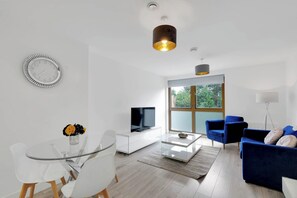Ein modernes Wohnzimmer mit einem Glastisch, einem Flachbildfernseher, einem blauen Sofa und einem großen Fenster mit Blick auf Grünflächen.